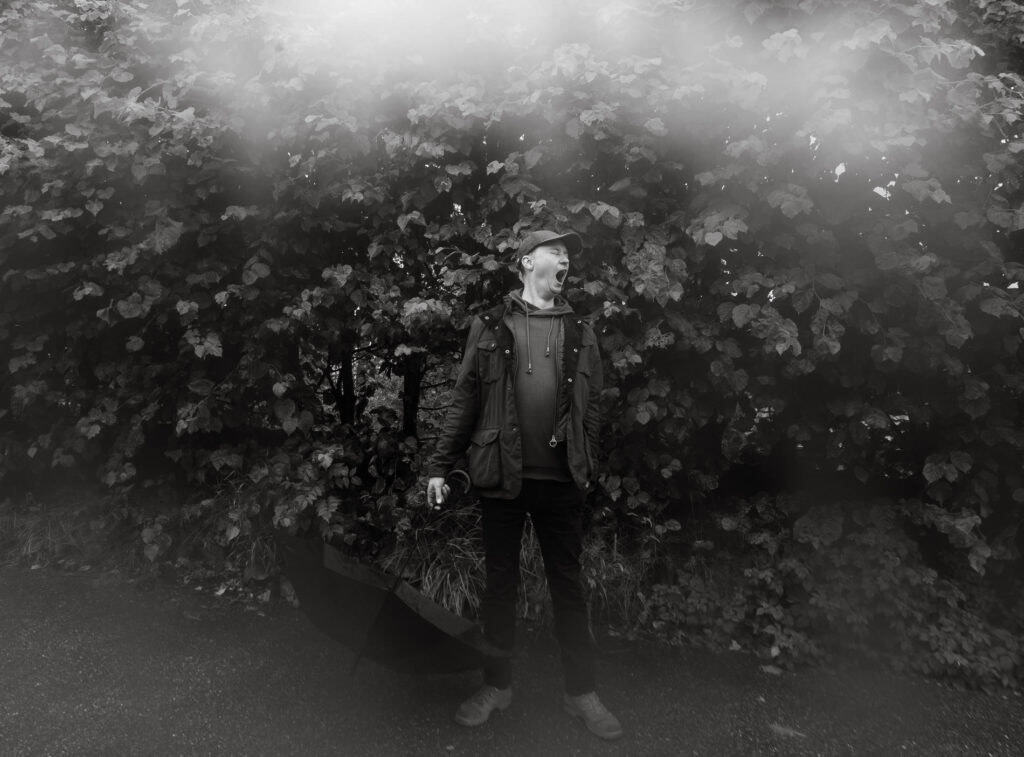 Dokumentarisk fotografi av en person som står foran buskads, med paraply i hånden.